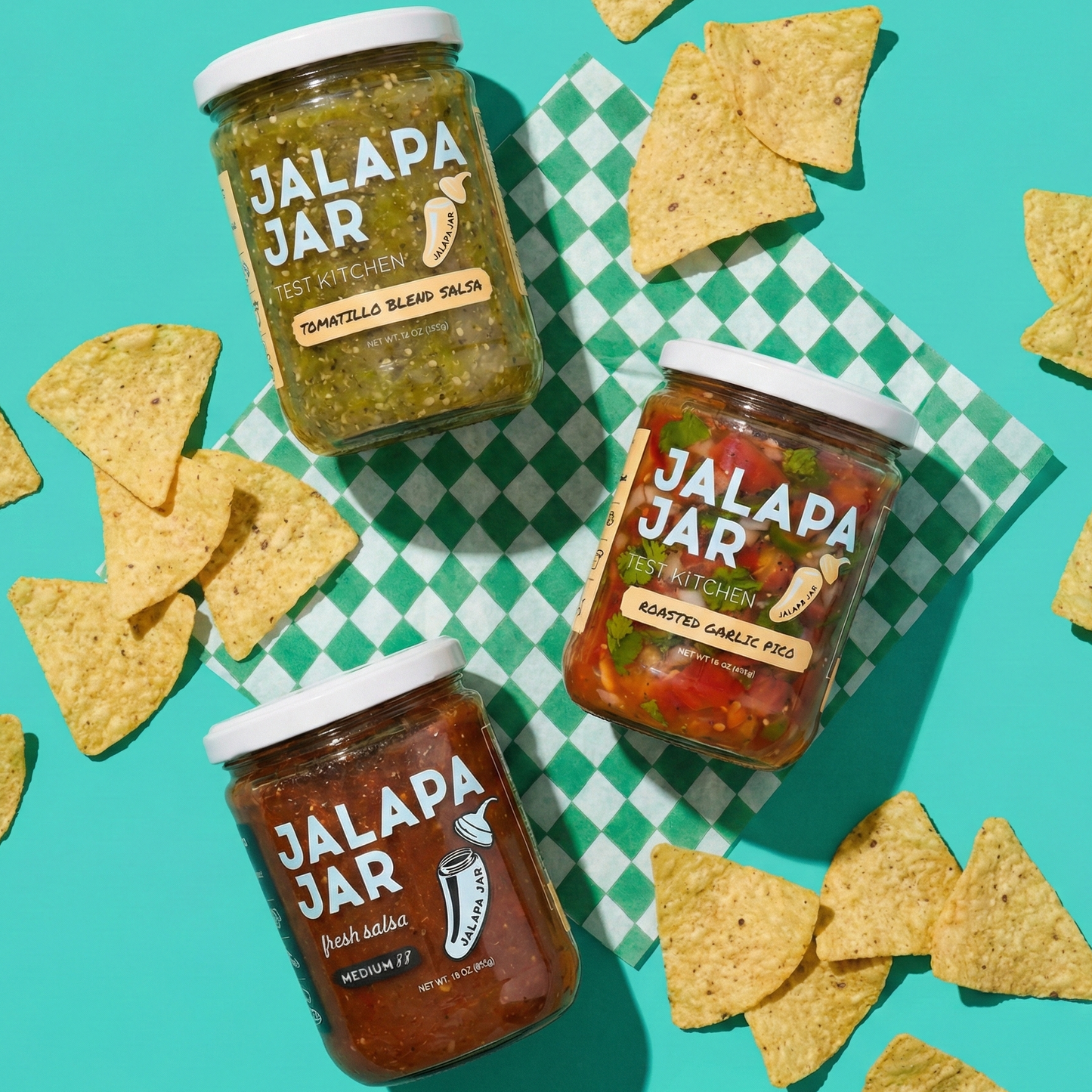 Three jars of Jalapa Jar salsa on a checkered tablecloth with tortilla chips around them.