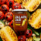 Jar of Jalapa Jar salsa surrounded by grilled pineapple and tomatoes on a wooden surface