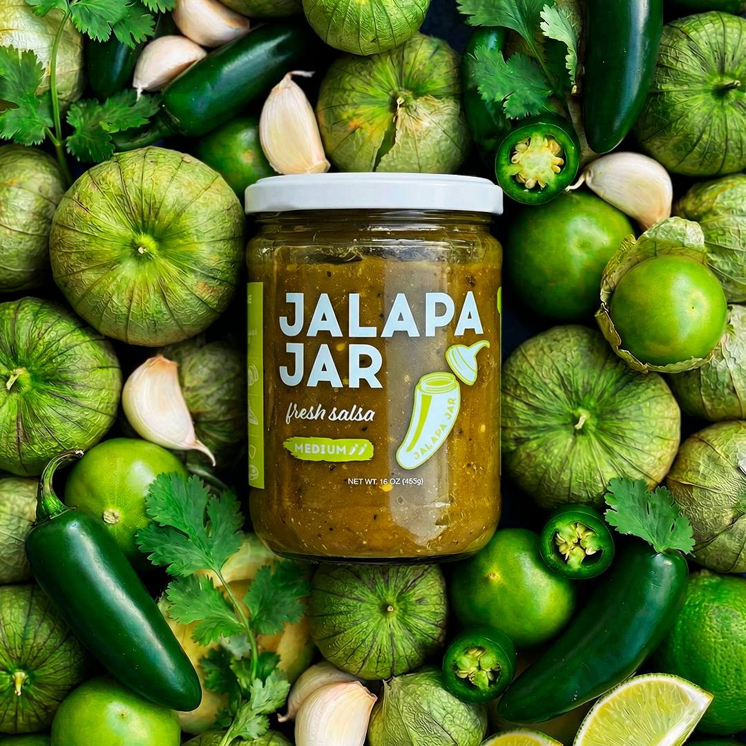 Jalapa Jar salsa surrounded by green peppers, jalapenos, and cilantro on a green background